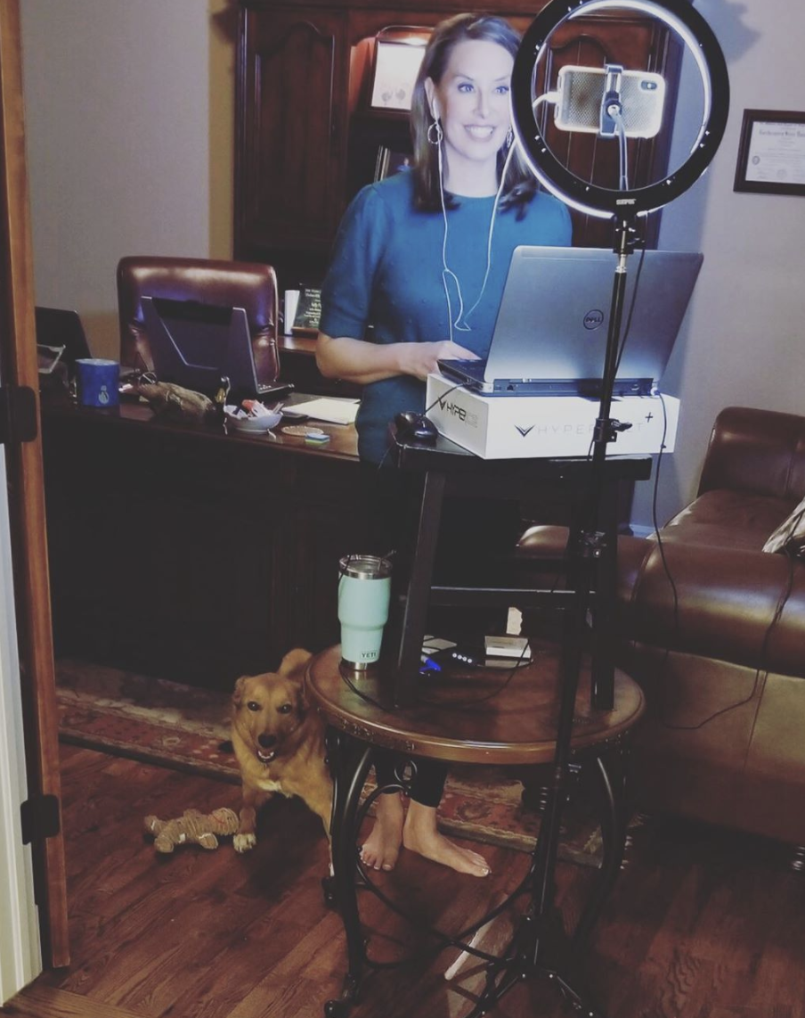 Image of Neile Jones standing in a room in her home using a cell phone on a lighted stand to broadcast the news from her home. She is barefoot and there is a dog at her feet.